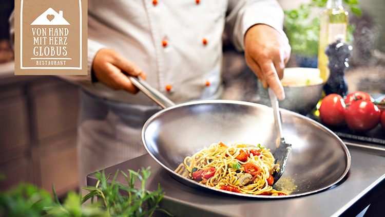 Ein Koch in weißer Jacke bereitet in einer Pfanne Spaghetti mit Tomaten zu, links Text: „Von Hand mit Herz GLOBUS Restaurant“.