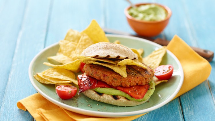 Burger mit gegrilltem Gemüse, Nachos, Tomatenhälften auf einem Teller, daneben eine Schale mit grünem Dip auf blauem Untergrund.