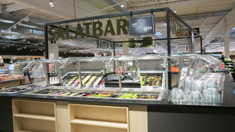 Eine Salatbar mit verschiedenen Salatzutaten, leeren durchsichtigen Schalen und einem Preisschild mit „Salate 0,89“.