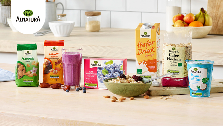 Mehrere Alnatura-Produkte, darunter Mandeln, Datteln, Haferflocken, Haferdrink, Joghurt und ein Beeren-Smoothie, auf einem Tisch.