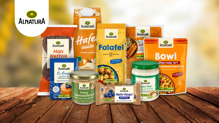 Produkte der Marke Alnatura, darunter Maiswaffeln, Bowl, Hafer Vanille und weitere, auf einer Holzoberfläche vor hellen Farben.