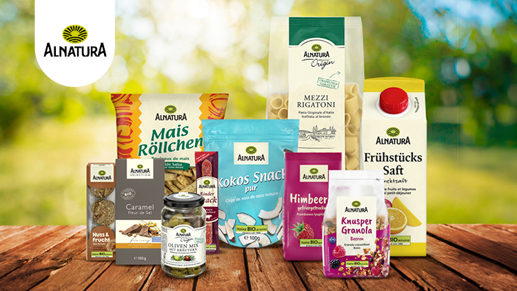 Alnatura-Produkte wie Snacks, Saft und Pasta auf einem Holztisch vor einer grünen, sonnigen Naturkulisse, Logo oben links.