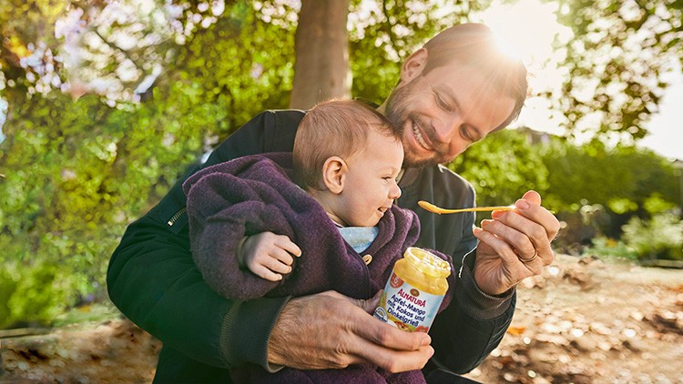 Erwachsene Person hält ein Baby und füttert es mit einem gelben Brei aus einem Gläschen der Marke Alete im Freien.