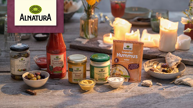 Alnatura-Produkte wie Oliven, Ketchup, Senf, vegane Mayo und Hummus stehen auf einem Holztisch, umgeben von Kerzen.