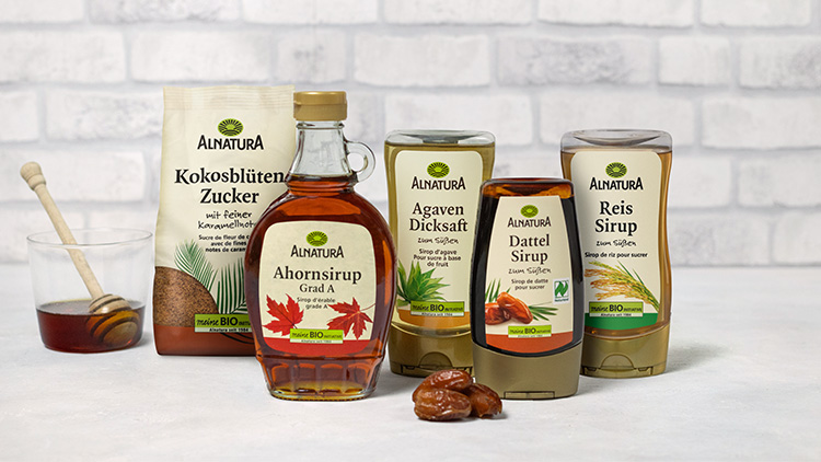 Fünf Alnatura Produkte, darunter Sirup, Zucker und Dicksaft, stehen neben einem Glas mit Honiglöffel auf hellem Untergrund.