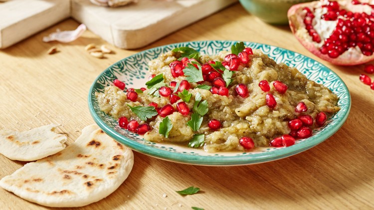 Püree mit Granatapfelkernen und Petersilie auf einem Teller, daneben Fladenbrot und Zutaten wie Knoblauch und Granatapfel.
