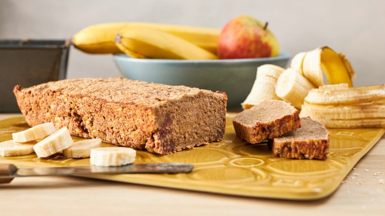 Ein Bananenbrot mit geschnittenen Bananenscheiben liegt auf einem gelben Teller, im Hintergrund sind Bananen und ein Apfel.