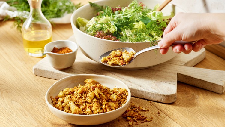 Eine Hand greift mit einem Löffel geröstete Gewürzstreusel aus einer Schale, daneben stehen Salat und eine Flasche mit Öl.