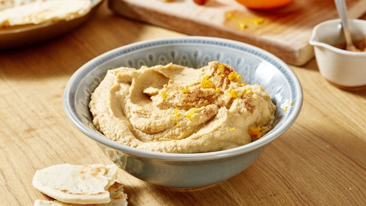 Schale mit cremigem Hummus, garniert mit Gewürzen und Zitronenzeste, daneben Fladenbrotstücke auf einem Holztisch.