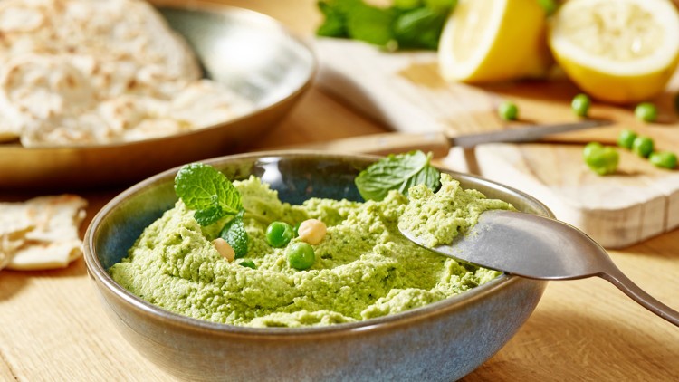 Grüne Creme mit Minzblättern, Kichererbsen und Erbsen in einer Schale, daneben angeschnittene Zitrone und Fladenbrot.