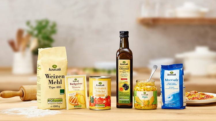 Mehrere Alnatura-Produkte, darunter Mehl, Backpulver, Tomatenstücke, Olivenöl, Mais und Meersalz, auf einer Holzarbeitsfläche.