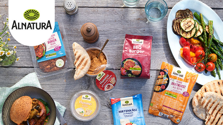 Holztisch mit Alnatura-Produkten wie Hummus, Burgerpatties, veganer Bratwurst, Grillgemüse, Fladenbrot und Blumenarrangement.