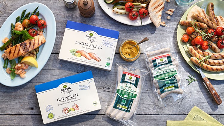 Tisch mit gegrilltem Gemüse, Würstchen, Lachs, Garnelen, Senf, Brot und Produkten von Alnatura wie Lachsfilets und Rostbratwürstchen.
