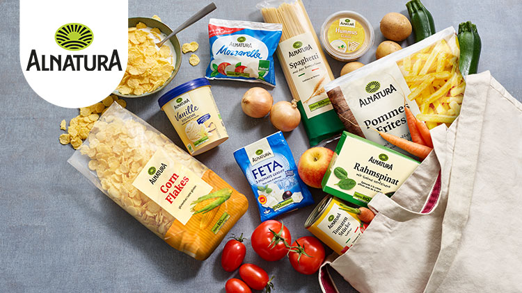 Mehrere Alnatura-Produkte wie Cornflakes, Spaghetti, Feta, Rahmspinat und Pommes frites liegen mit Gemüse auf einer grauen Fläche.