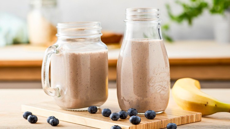 Zwei Gläser mit Bananen-Heidelbeer-Smoothie stehen auf einem Holzbrett, daneben liegen eine Banane und mehrere Heidelbeeren.