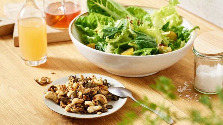 Eine Schüssel mit frischem grünen Salat, ein Teller mit Cashewkernen und Kürbiskernen, Honigglas und Essigkaraffe auf Holztisch.