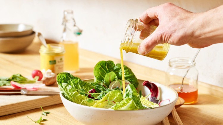 Grüne Salatblätter in einer weißen Schüssel werden mit gelbem Dressing aus einem Glas übergossen, im Hintergrund Zutaten.