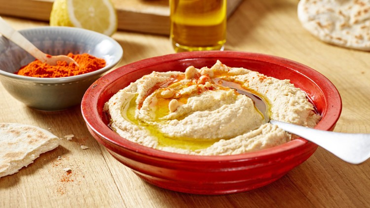 Roter Teller mit Hummus, darüber Öl, Kirchererbsen und Paprikapulver, daneben Fladenbrot, Gewürzschale und ein Glas Öl.