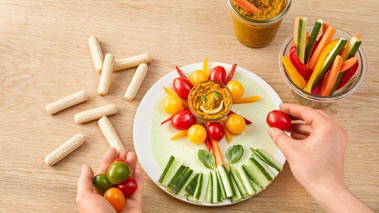 Teller mit Gemüse in Sonnenform und Dip in der Mitte, daneben Gemüsesticks und Brotsticks auf Holzoberfläche.