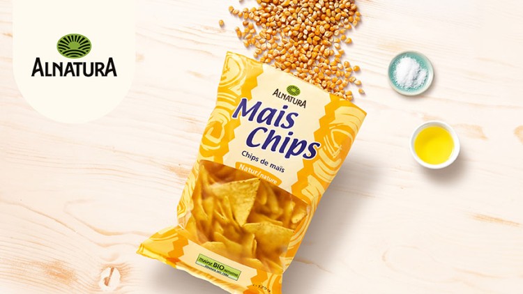 Eine geöffnete Packung Alnatura Mais Chips liegt neben Maiskörnern, einem Schälchen Salz und einer Schale mit gelber Flüssigkeit.