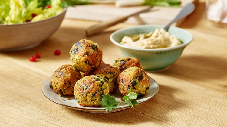 Sechs gebratene Falafel auf einem Teller mit frischem Koriander, daneben eine Schale Hummus und ein Salat im Hintergrund.