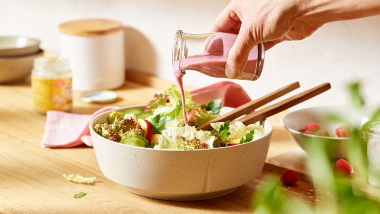 Eine Hand gießt pinkes Dressing aus einer kleinen Glasflasche über einen frischen Salat in einer hellen Schüssel auf Holzuntergrund.