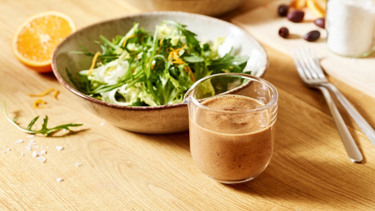 Salat mit grünem Blattgemüse in einer Schale, daneben ein Glas Dressing, Orangenhälfte, Oliven und Besteck auf einem Holztisch.
