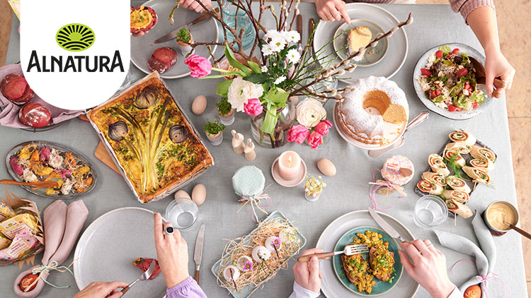 Frühlingshaft gedeckter Tisch mit Alnatura-Logo, Speisen wie Rote-Bete-Brötchen, Gemüsequiche, Gugelhupf, Wraps und frischen Blumen.