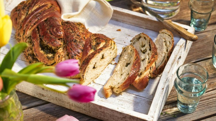 Ein Holztablett mit aufgeschnittenem Kräuterbrot, daneben ein Messer und Gläser, im Vordergrund gelbe und violette Tulpen.