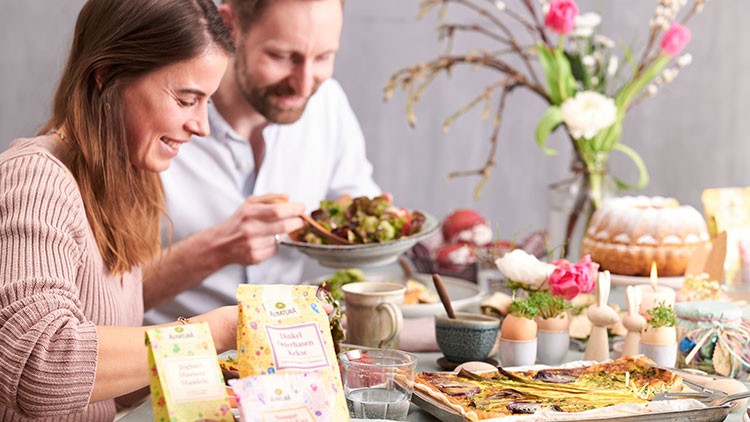 Ein Tisch mit gedecktem Geschirr, Blumen, Osterdekoration, Speisen wie Quiche und Kuchen sowie Produkten von Sonnentor.