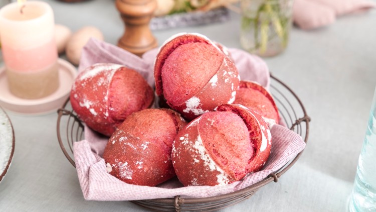 Rote Brötchen mit Mehl bestäubt liegen in einem Metallkorb auf einem Stofftuch, im Hintergrund Tischdekoration.