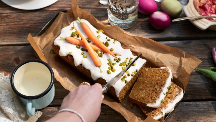 Ein Karottenkuchen mit weißem Guss, Pistazien und kleinen Karotten wird auf Backpapier liegend angeschnitten.