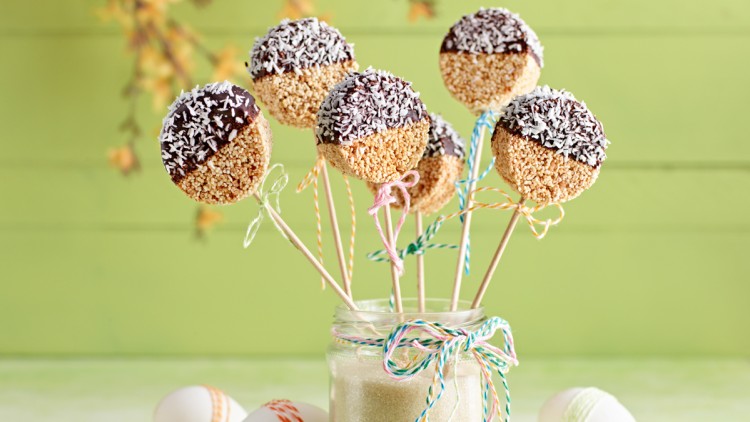 Bunte Cake-Pops mit Schokolade und Kokosraspeln stecken in einem Glas mit Zuckersand, umgeben von dekorierten Ostereiern.