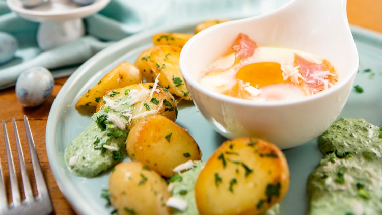 Gebratene Kartoffeln mit Kräutern und grüner Soße neben einem kleinen Schälchen mit Spiegelei und Schinken auf einem Teller.