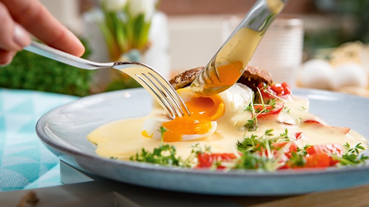 Ein pochiertes Ei mit flüssigem Eigelb auf Sauce Hollandaise, garniert mit Kresse und Tomaten, wird mit Messer und Gabel geschnitten.