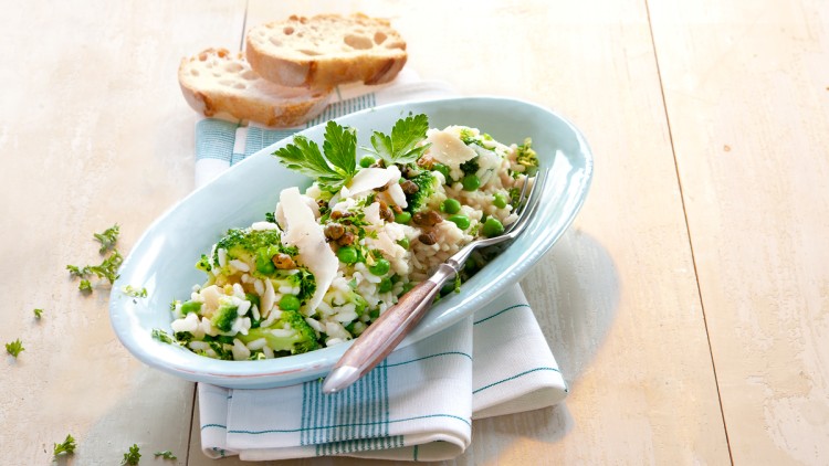 Cremiges Risotto mit Erbsen, Brokkoli und Kräutern in einer ovalen Schale, garniert mit Parmesan, daneben zwei Scheiben Baguette.