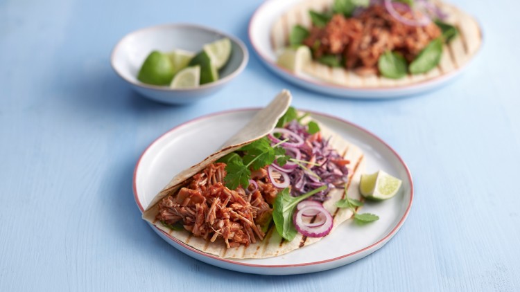 Ein gefüllter Taco mit Jackfruit, Rotkohl, roten Zwiebeln, Blattgrün und Koriander liegt auf einem Teller mit Limettenstücken.