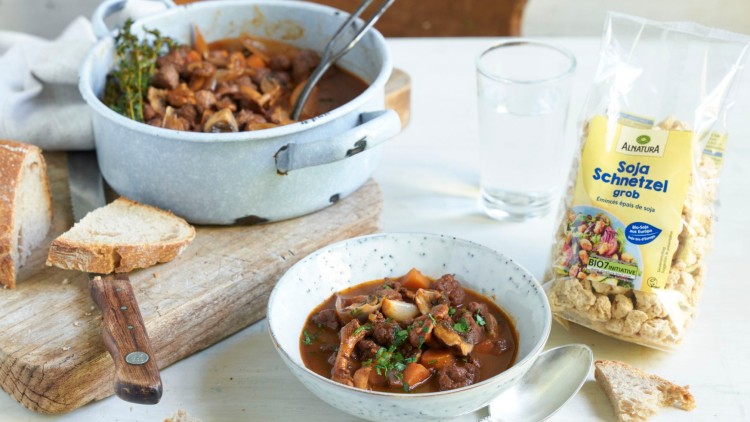Ein Teller mit Soja-Gulasch steht vor einem Topf, daneben liegen Brotstücke und eine Packung Alnatura Soja-Schnetzel grob.