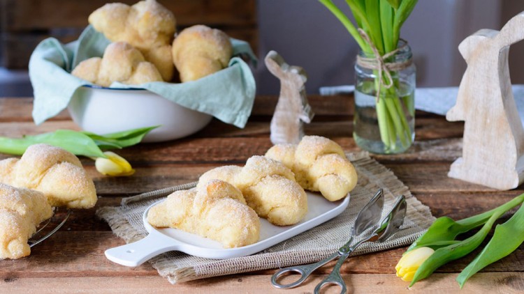 Mehrere gezuckerte Hefezöpfe auf einer weißen Platte, daneben Tulpen, Holzhasen und eine Schale mit weiteren Zöpfen.