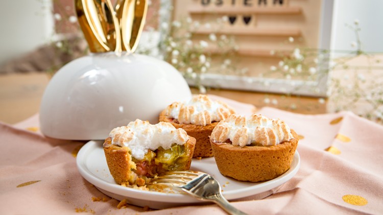 Drei kleine Kuchen mit Baiserhaube auf weißem Teller, einer angeschnitten, goldener Löffel daneben, Hintergrund mit Osterdekoration.