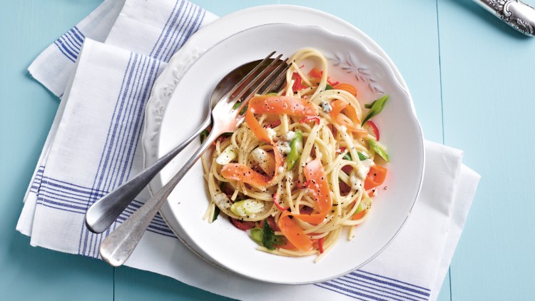 Ein Teller mit Spaghetti, Karottenstreifen, grünen Zwiebeln, Kräutern und Pfeffer auf weißem Stoff mit blauem Streifenmuster.