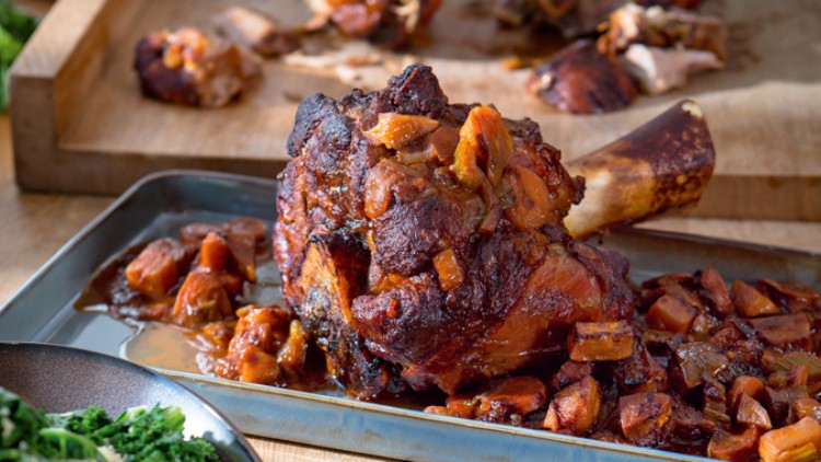 Eine gebratene Schweinshaxe mit Gemüsewürfeln in Soße liegt auf einer Metallplatte, im Hintergrund ein Holzbrett.