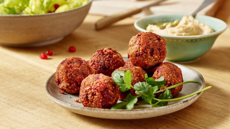 Rote Falafelbällchen mit frischem Koriander auf einem Teller, daneben Hummus in einer Schale und ein grüner Salat.