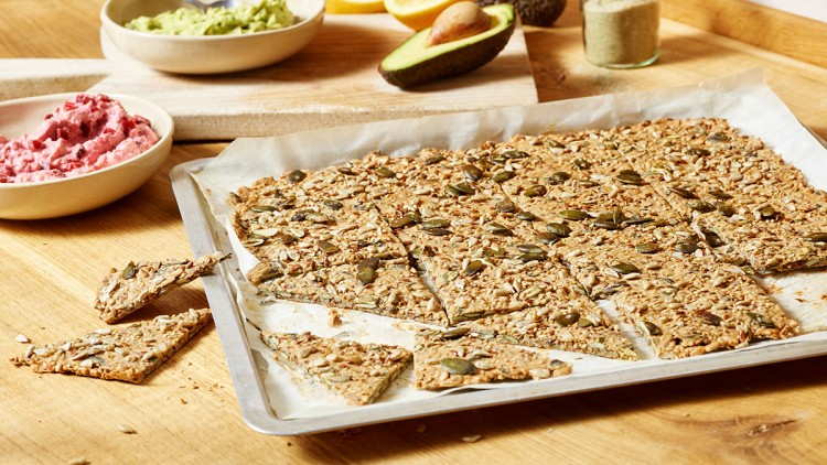 Knuspriges Knäckebrot mit Körnern auf einem Backblech, daneben Schalen mit grüner Avocadocreme und pinkem Aufstrich.
