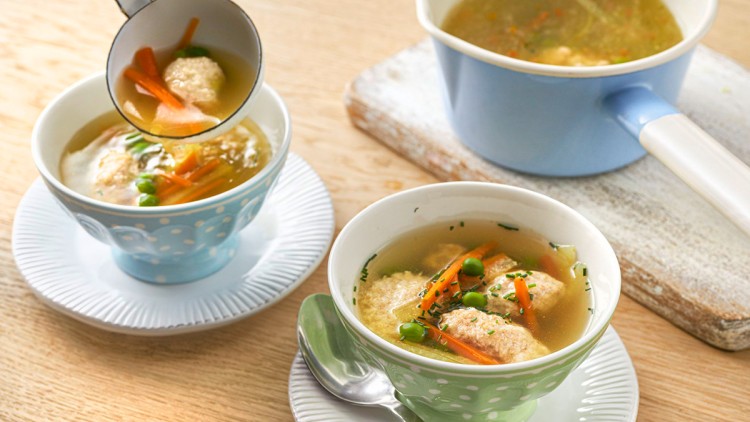 Zwei gepunktete Schalen mit klarer Suppe, Gemüse, Klößen und Löffeln stehen auf Tellern, daneben ein Topf und eine Suppenkelle.