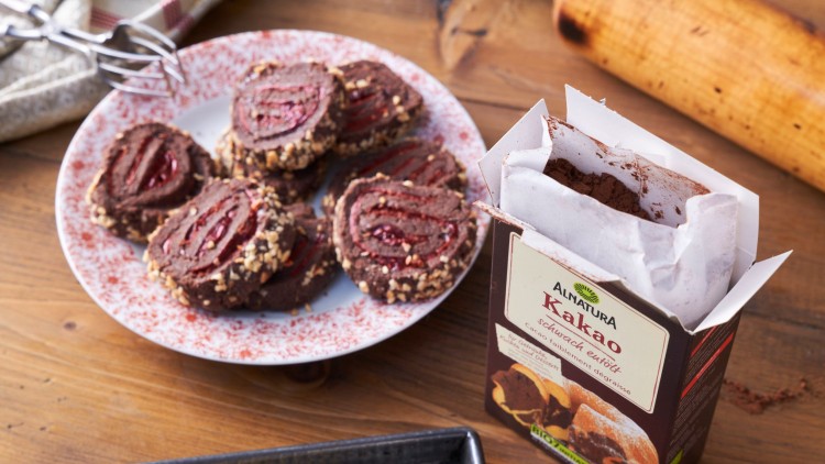 Ein Teller mit Schokokeksen, gefüllt mit roter Marmelade und gehackten Nüssen, steht neben einer geöffneten Kakaoverpackung.