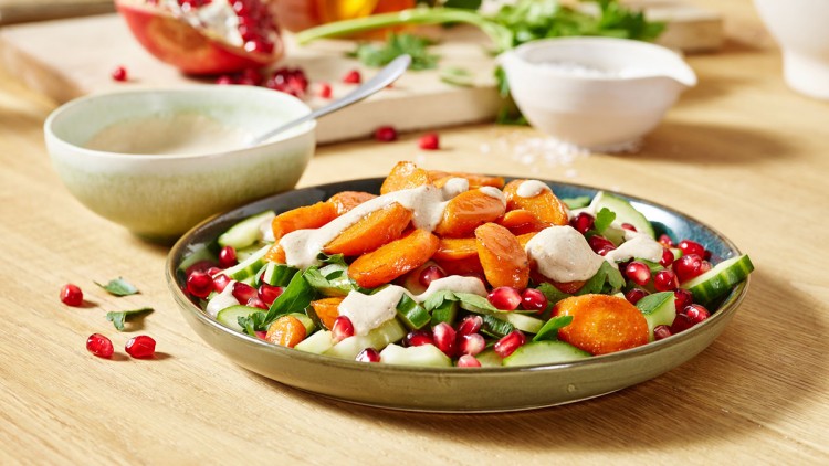 Teller mit Salat aus Gurken, Granatapfelkernen, karamellisierten Karotten und Soße, daneben eine Schale und Kräuter.