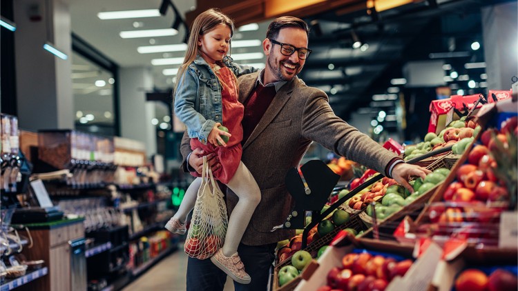 Erwachsener hält ein Kind auf dem Arm, beide betrachten Obst in einem Supermarkt, das Kind hält ein Netz mit Einkäufen.