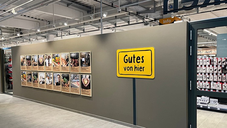 Graue Wand mit gelbem Schild und schwarzer Aufschrift „Gutes von hier“, daneben zwölf Bilder mit Beschreibungen und Produkten.