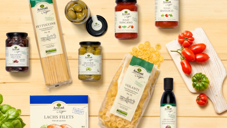 Mehrere Alnatura-Produkte, darunter Nudeln, Saucen, Oliven und Lachsfilets, arrangiert auf hellem Holz mit frischen Kräutern und Tomaten.